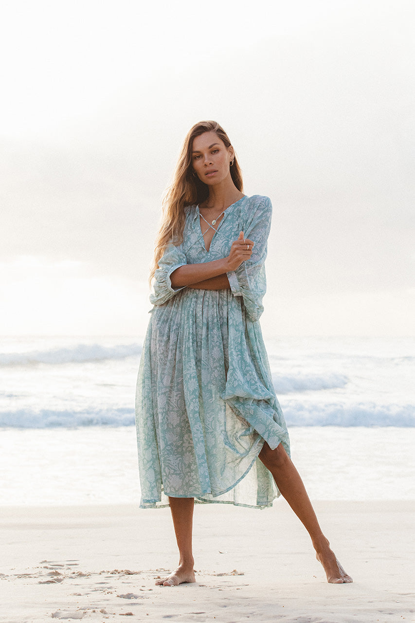 Woman wearing Daughters of India Kyra Midi Dress in Jade, a floral blue-green chiffon dress, standing on a beach