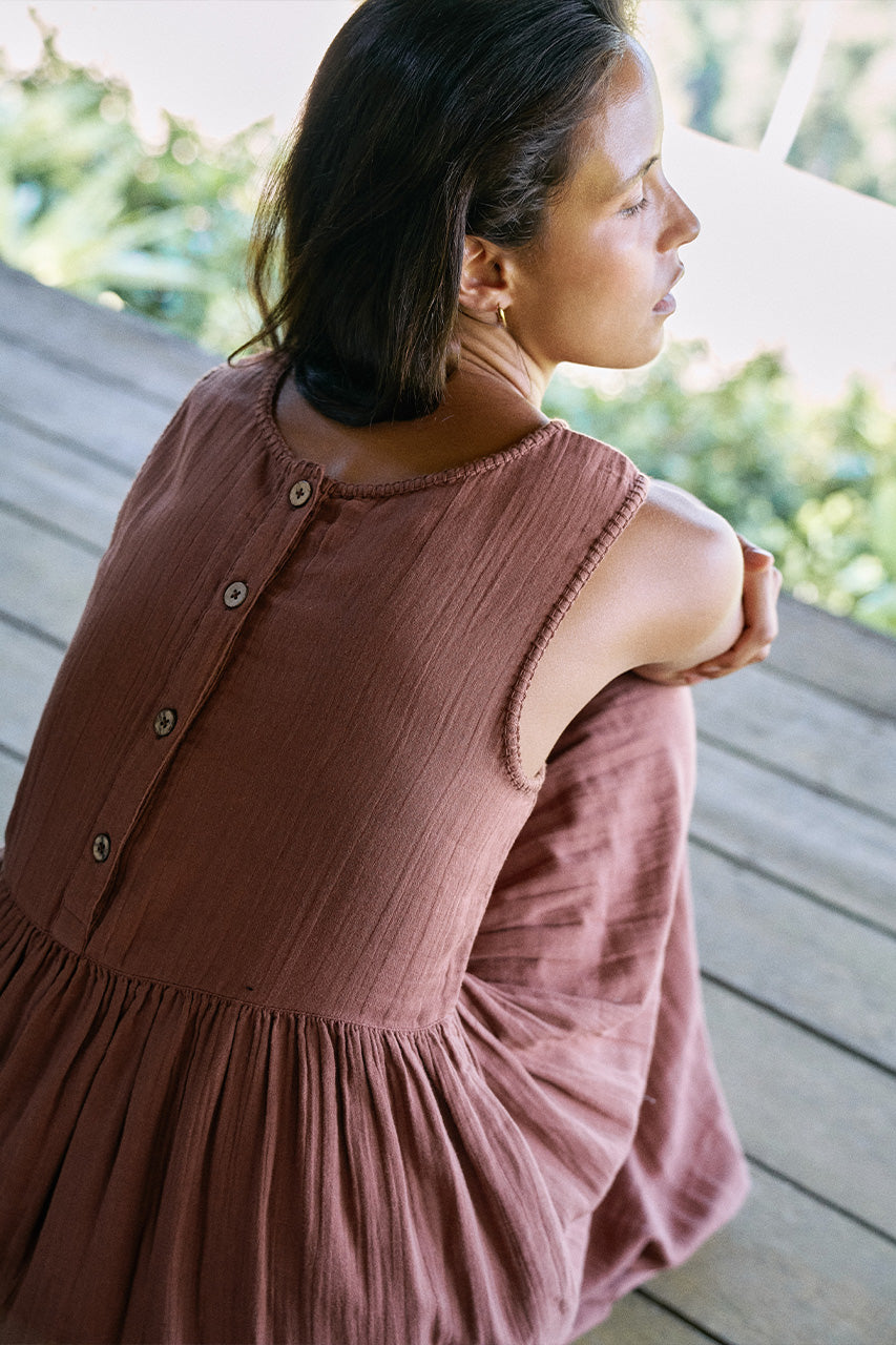 Nadi Dress Chocolate — close-up back view seated on wooden deck, back button detail and gathered waist, chocolate brown double gauze cotton, tropical greenery background | Daughters of India