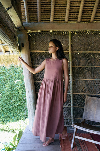 Nadi Dress Chocolate — full length front view standing in bamboo hut doorway looking out, chocolate brown double gauze cotton midi dress with gathered waist, woven bamboo walls | Daughters of India