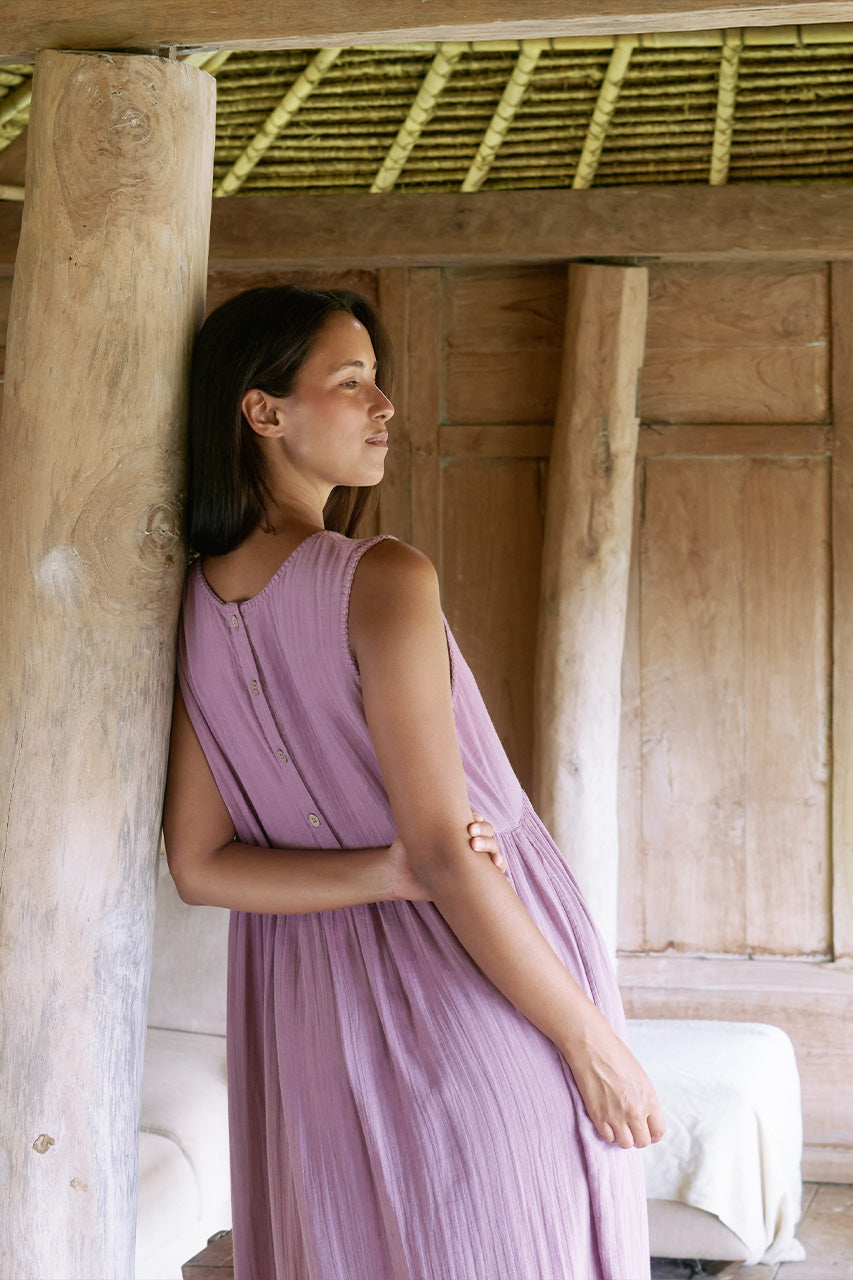 Nadi Dress Mauve — three-quarter view leaning against wooden post, mauve double gauze cotton midi dress with wooden button front, rustic wooden beams and thatched roof | Daughters of India