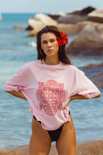 Shanti Tee Pink — front view at beach rocks, pink oversized cotton tee with red hand-printed Daughters of India mandala logo, red hibiscus flower in hair, ocean rocks backdrop | Daughters of India